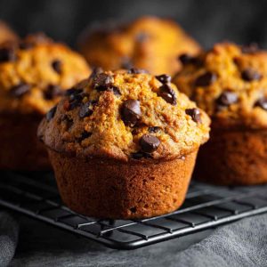 Freshly baked pumpkin chocolate chip muffins with golden tops and gooey chocolate chips, served on a rustic tray with fall leaves and a warm drink.