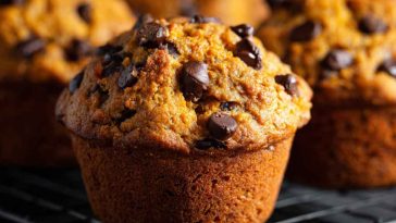 Freshly baked pumpkin chocolate chip muffins with golden tops and gooey chocolate chips, served on a rustic tray with fall leaves and a warm drink.