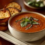 A bowl of creamy roasted tomato basil soup garnished with fresh basil leaves and served with crusty bread on a rustic wooden table.