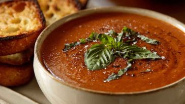 A bowl of creamy roasted tomato basil soup garnished with fresh basil leaves and served with crusty bread on a rustic wooden table.
