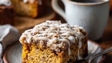 A slice of moist pumpkin coffee cake topped with cinnamon streusel and drizzled with glaze, served on a rustic plate with fall decor.
