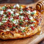 A golden pizza topped with spicy sausage, crumbled feta, and a drizzle of honey, served on a rustic board with chili flakes and fresh herbs.
