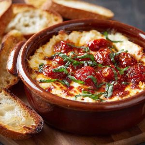 Three-Cheese Tomato Bruschetta Dip served warm in a skillet, topped with melted mozzarella, parmesan, and cream cheese, fresh tomatoes, basil, and toasted baguette slices.