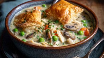 High protein chicken pot pie soup served in a bowl with shredded chicken, peas, carrots, and creamy broth, garnished with fresh herbs and a puff pastry topper.