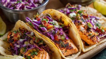 Air fryer salmon tacos topped with cabbage slaw, lime wedges, and cilantro on a serving board.