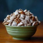 Caramel apple puppy chow coated in powdered sugar and caramel drizzle, mixed with dried apple bits and served in a festive bowl.