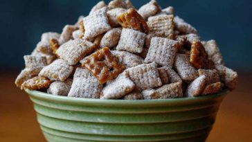 Caramel apple puppy chow coated in powdered sugar and caramel drizzle, mixed with dried apple bits and served in a festive bowl.