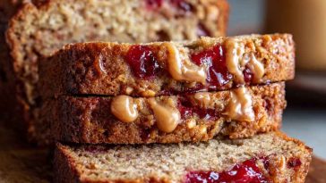 Sliced peanut butter & jelly banana bread with swirls of jam and creamy peanut butter, served on a rustic board with fresh bananas and a jar of jelly.