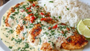 A plate of Mexican-style chicken smothered in creamy cheese sauce, garnished with chopped cilantro and served with rice and sautéed peppers.