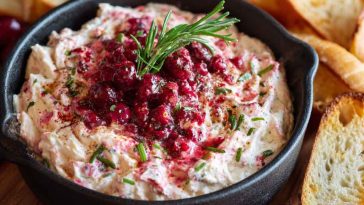 A warm cranberry and cream cheese dip served in a baking dish, swirled with ruby-red cranberry sauce and topped with chopped pecans and fresh rosemary.