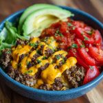 A deconstructed cheeseburger bowl with seasoned ground beef, shredded lettuce, cherry tomatoes, pickles, cheddar cheese, and a drizzle of burger sauce.