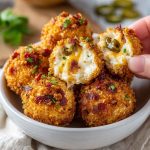 Crispy bacon jalapeño popper cheese balls served on a platter with dipping sauce, garnished with chopped herbs and jalapeño slices.