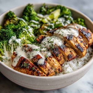 A bowl filled with grilled chicken slices, steamed broccoli, and rice, drizzled with creamy garlic sauce and garnished with parsley and lemon wedges.