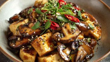 A sizzling skillet of mushroom and tofu stir-fry with bell peppers, scallions, and sesame seeds, coated in a glossy soy-ginger sauce and served over steamed rice.