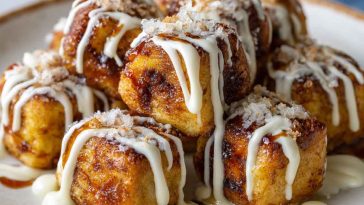 A plate of golden cinnamon roll French toast bites drizzled with icing and dusted with powdered sugar, served with fresh berries and maple syrup.
