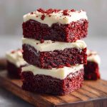 Fudgy red velvet brownies topped with swirls of cream cheese frosting, garnished with red sprinkles on a white plate.