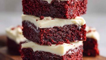 Fudgy red velvet brownies topped with swirls of cream cheese frosting, garnished with red sprinkles on a white plate.