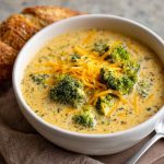 A steaming bowl of creamy broccoli cheddar soup topped with shredded cheese and cracked pepper, served with crusty bread on a rustic wooden table.