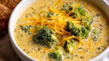 A steaming bowl of creamy broccoli cheddar soup topped with shredded cheese and cracked pepper, served with crusty bread on a rustic wooden table.