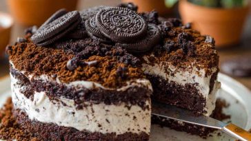 A layered dessert of crushed OREO cookies and creamy pudding mixture in a glass dish, topped with gummy worms and served with a spoon.