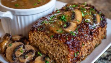 A thick slice of meatloaf topped with rich mushroom gravy, served with mashed potatoes and green beans on a rustic dinner plate.