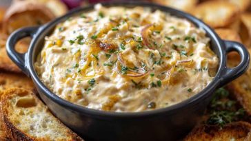 A bubbling dish of smoked gouda and onion dip with golden edges, topped with caramelized onions and served with toasted baguette slices.