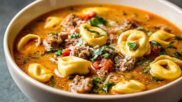 A steaming bowl of sausage and tortellini soup filled with cheesy pasta, crumbled sausage, spinach, and a creamy tomato broth, served with a slice of rustic bread.
