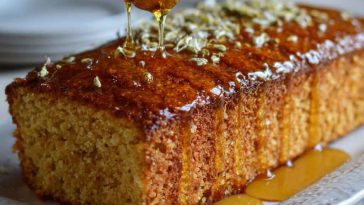 A golden slice of Greek honey cake topped with crushed walnuts and a drizzle of honey, served on a rustic ceramic plate with a sprig of thyme.