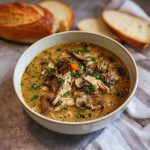 A bowl of creamy rotisserie chicken and mushroom soup garnished with fresh thyme and cracked black pepper, served with crusty bread on the side.