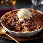 A golden-brown pecan pie dump cake fresh from the oven, topped with crunchy pecans and gooey caramel filling, served in a rustic baking dish with a scoop of vanilla ice cream.