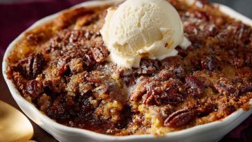 A golden-brown pecan pie dump cake fresh from the oven, topped with crunchy pecans and gooey caramel filling, served in a rustic baking dish with a scoop of vanilla ice cream.