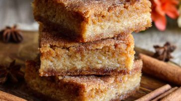 A batch of cinnamon sugar blondies cut into squares, with golden edges and a crackly cinnamon topping, served on a rustic tray.