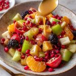A vibrant bowl of winter fruit salad featuring pomegranate seeds, citrus slices, apples, and pears, drizzled with cinnamon vanilla dressing.
