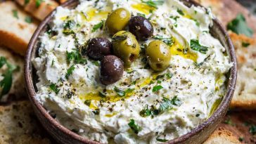 A bowl of creamy feta and green olive dip topped with chopped herbs and olive oil, surrounded by pita chips and fresh veggies.