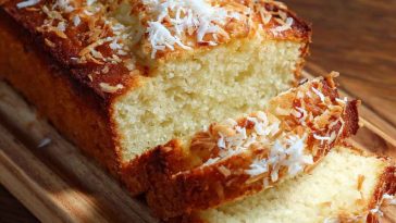 Moist coconut loaf cake topped with shredded coconut, sliced and served on a tropical-inspired platter.