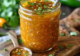 A jar of mango habanero honey garlic sauce with visible chili flakes, surrounded by fresh mango slices, garlic cloves, and habanero peppers.