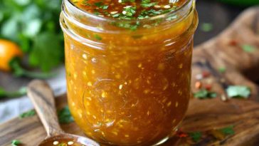 A jar of mango habanero honey garlic sauce with visible chili flakes, surrounded by fresh mango slices, garlic cloves, and habanero peppers.