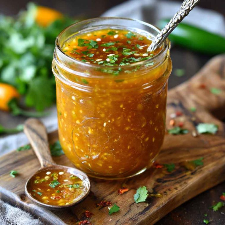 A jar of mango habanero honey garlic sauce with visible chili flakes, surrounded by fresh mango slices, garlic cloves, and habanero peppers.