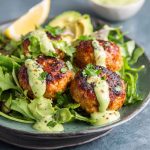 Salmon meatballs drizzled with creamy avocado sauce, served with fresh herbs and lemon wedges.