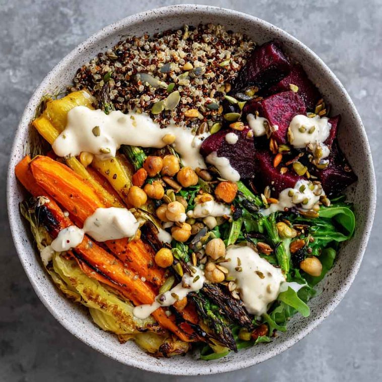 Colorful plant-based bowl filled with roasted vegetables, grains, greens, and drizzled with creamy tahini yogurt sauce.