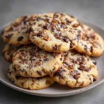 Golden shortbread cookies studded with chocolate chips and crunchy toffee bits, stacked on a plate with crumbs scattered around.