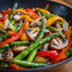 A skillet filled with colorful sautéed vegetables including bell peppers, zucchini, carrots, and broccoli, garnished with fresh herbs.