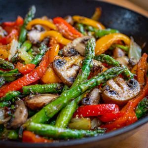 A skillet filled with colorful sautéed vegetables including bell peppers, zucchini, carrots, and broccoli, garnished with fresh herbs.