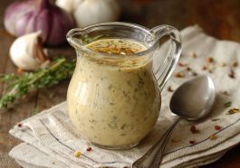 A glass jar of creamy parmesan pepper dressing with visible cracked black pepper, surrounded by fresh salad greens and lemon wedges.