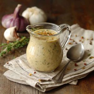 A glass jar of creamy parmesan pepper dressing with visible cracked black pepper, surrounded by fresh salad greens and lemon wedges.