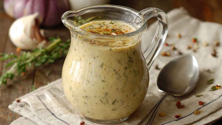 A glass jar of creamy parmesan pepper dressing with visible cracked black pepper, surrounded by fresh salad greens and lemon wedges.
