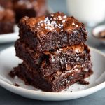 A stack of fudgy zero sugar brownies made with Greek yogurt, topped with a dusting of cocoa powder and fresh raspberries.