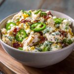 A bowl of jalapeno popper chicken salad with shredded chicken, diced jalapeños, crispy bacon, and melted cheese, garnished with fresh herbs.