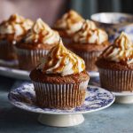 A tray of sticky toffee cupcakes topped with glossy toffee sauce and a swirl of whipped cream, garnished with chopped dates.