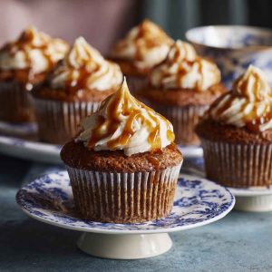 A tray of sticky toffee cupcakes topped with glossy toffee sauce and a swirl of whipped cream, garnished with chopped dates.
